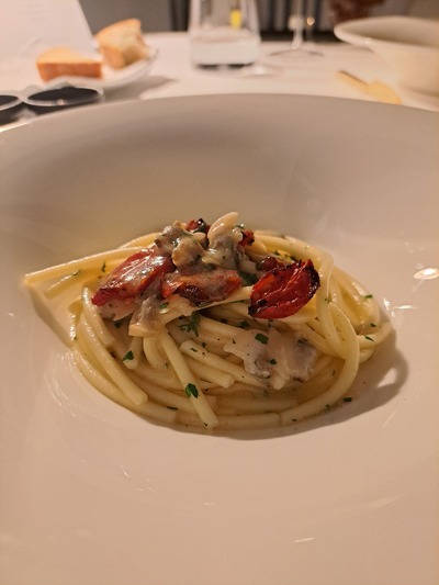 Spaghetti affumicati con vongole e datterini arrostiti. Uno dei più grandi classici della cucina di mare italiana rivisitato da chef Uliassi in chiave moderna. Un piatto che fa della semplicità concettuale la sua forza, esaltando consistenze e sapori di ciascun ingrediente.