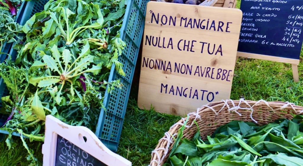Una cassetta di plastica contenente erbe di campo e una cesta di legno contenente aglio orsino sono posizionate a terra sull'erba, vicino a una tavolozza di legno dove è incisa la suggestiva frase "Non mangiare nulla che tua nonna non avrebbe mangiato."