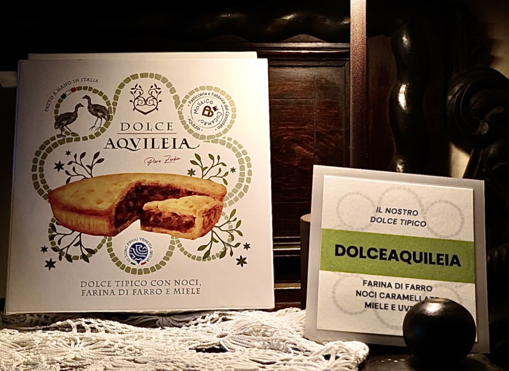 Confezione del dolce Aquileia, esposta su un antico mobile, che riporta gli ingredienti della torta e l'indicazione che si tratta di un dolce realizzato a mano dalla pasticceria Mosaico.