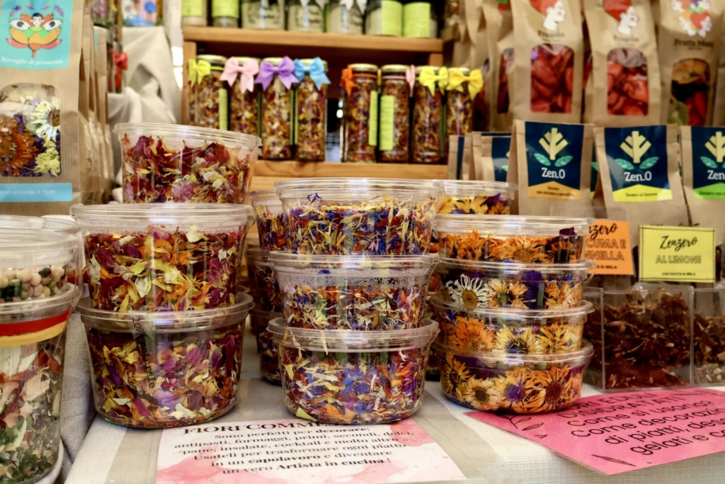 Diverse tipologie di variopinti fiori edibili secchi posti nelle confezioni di plastica sigillate, sullo sfondo compaiono altri prodotti esiccati a base di frutta e fiori.