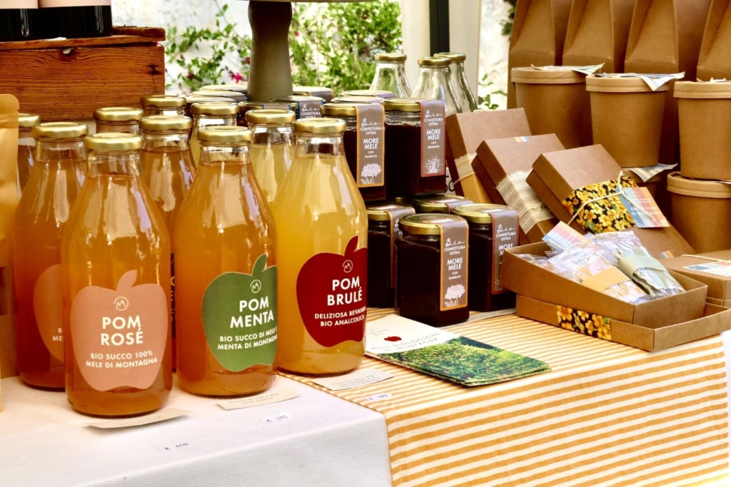 tre diverse tipologie di succo di mela biologico in bottiglie di vetro, vicino a dei barattoli di marmellata artigianale alle mele e more e a delle curate confezioni di infusi misti per tisane, sul tavolo è posizionato anche anche il depliant dell'azienda produttrice Naturalpina.