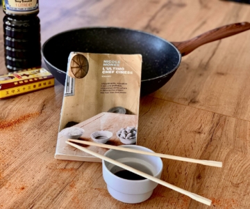 Su un tavolo di legno dove sono sparse un po' di spezie in polvere, sono appoggiati: un wok, una bottiglia di salsa di soia, una ciotolina con delle bacchette sopra e al centro il romanzo L'ultimo Chef Cinese di Nicole Mones.