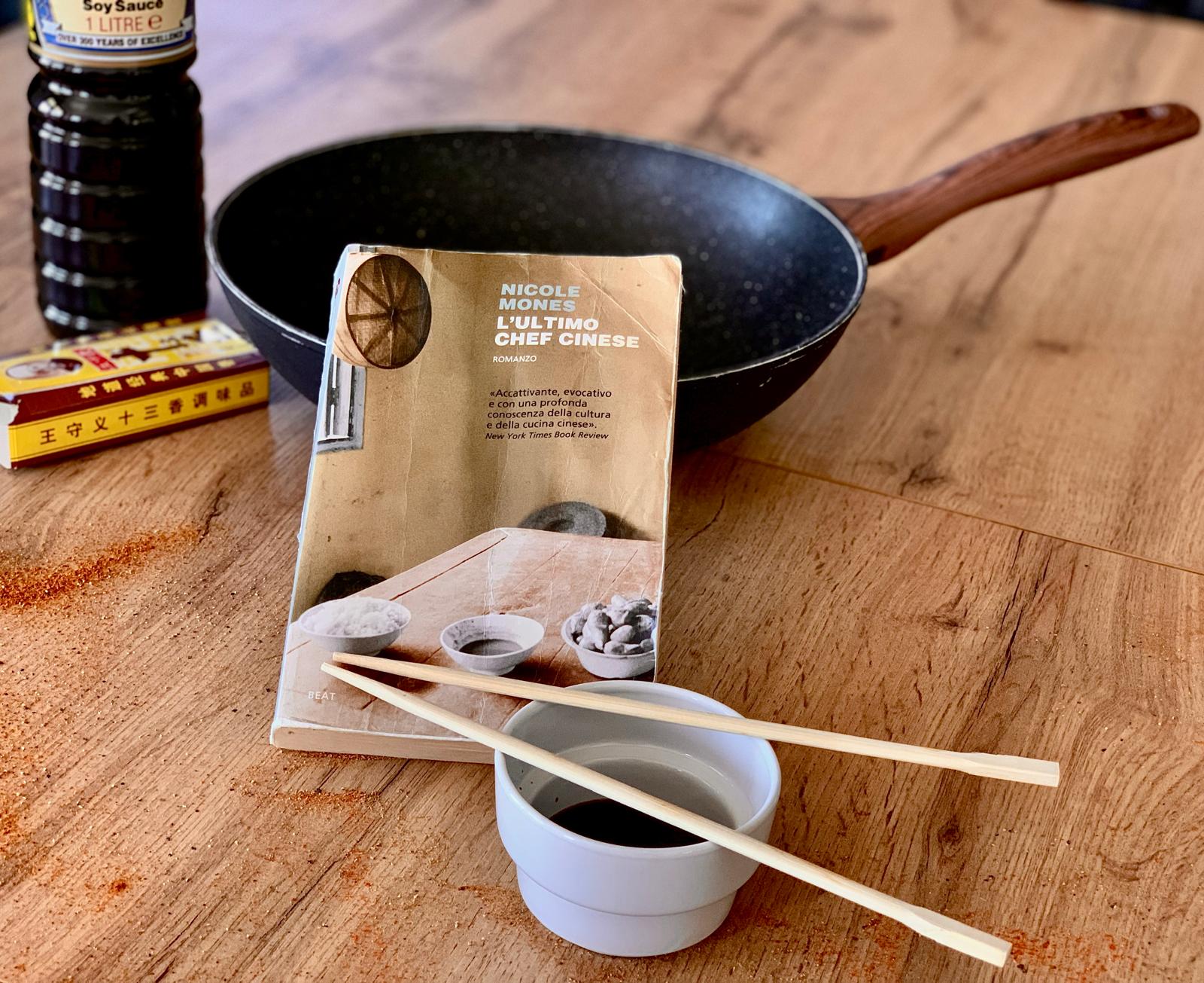 Su un tavolo di legno dove sono sparse un po' di spezie in polvere, sono appoggiati: un wok, una bottiglia di salsa di soia, una ciotolina con delle bacchette sopra e al centro il romanzo L'ultimo Chef Cinese di Nicole Mones.